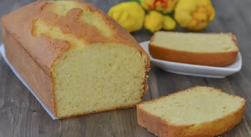 Un soffice plumcake al limone, appena sfornato, con una fetta su un piattino bianco e fiori gialli sullo sfondo.