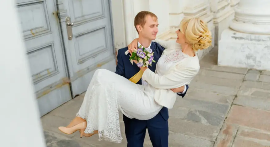Pan młody niesie pannę młodą na rękach. Zastanawiasz się nad skutkami spóźnienia się z rezerwacją fotografa? Dlaczego 18 miesięcy to minimum?