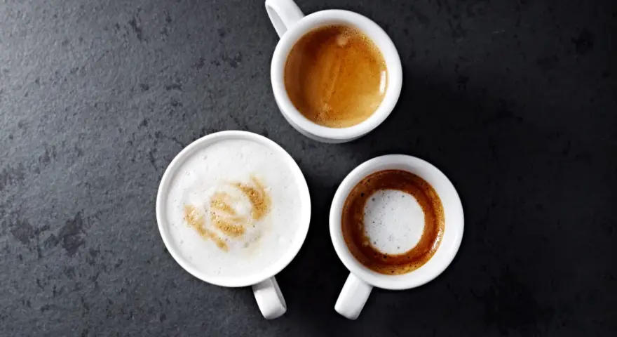 Trzy białe filiżanki z kawą: espresso, latte macchiato i kawą z pianką na ciemnym tle.