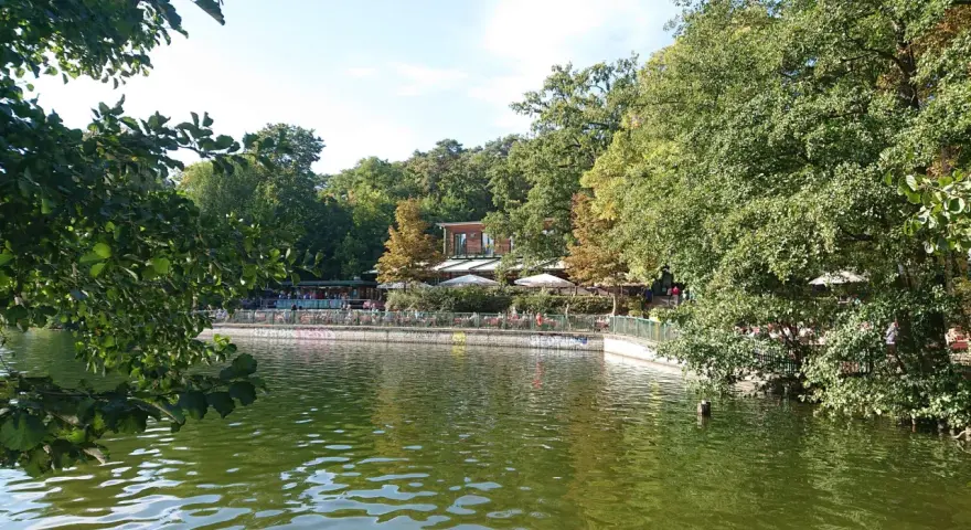 Grüne Bäume säumen den Schlachtensee Berlin, wo Menschen auf der Terrasse eines Restaurants sitzen.