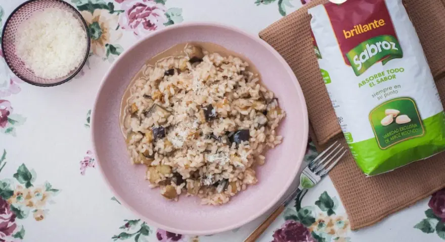 Arroz meloso con berenjenas en plato rosa, acompañado de queso rallado y bolsa de arroz Sabroz.