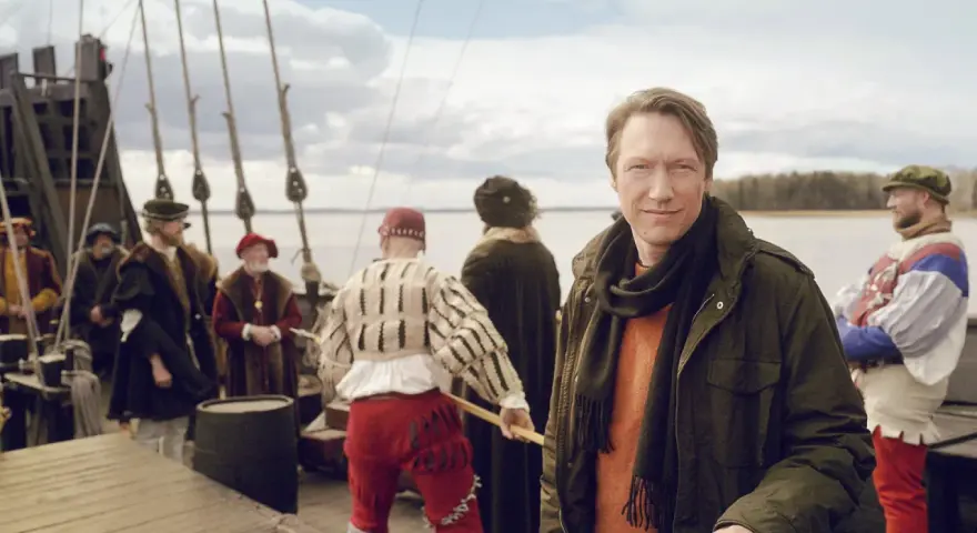 Man i modern jacka på en historisk båt med folk i tidsenliga kläder. En påminnelse om Sveriges långa historia.