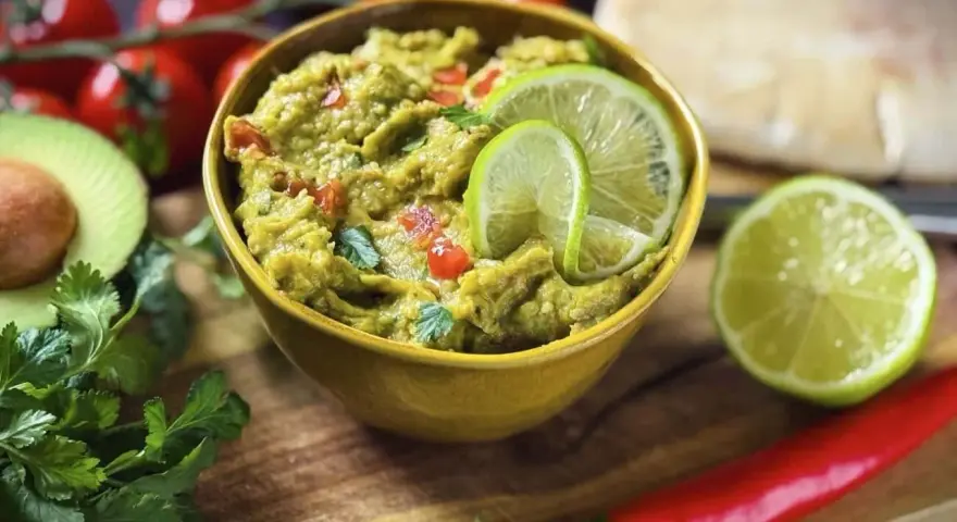Frisches Guacamole-Rezept in einer gelben Schüssel, garniert mit Limettenscheiben. Daneben liegen Avocado, Koriander, Tomaten und Chili.