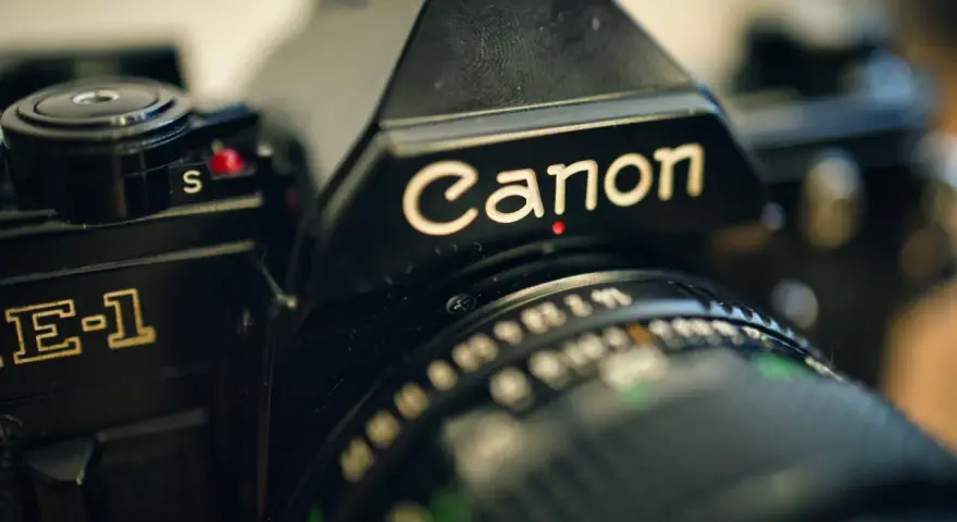 Zbliżenie na czarną lustrzankę Canon AE-1. Widoczny napis "Canon" i fragment obiektywu. Idealny do połączenia z aparatem.