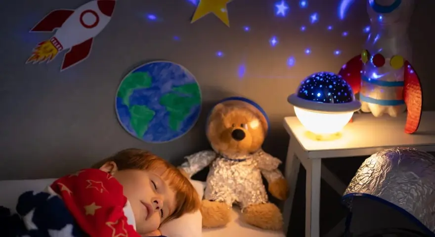 Śpiące dziecko pod kocem w gwiazdki, obok pluszowy miś w stroju astronauty. Na ścianie rakieta i Ziemia, a na suficie projektor dla dzieci na sufit wyświetla gwiazdy.