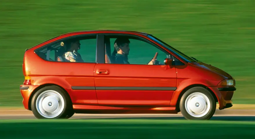 Czerwony samochód elektryczny BMW E1 z lat 90. jadący po zielonym tle.