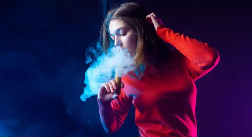 Joven exhala humo de un vaper, rodeada de luces azules y moradas.