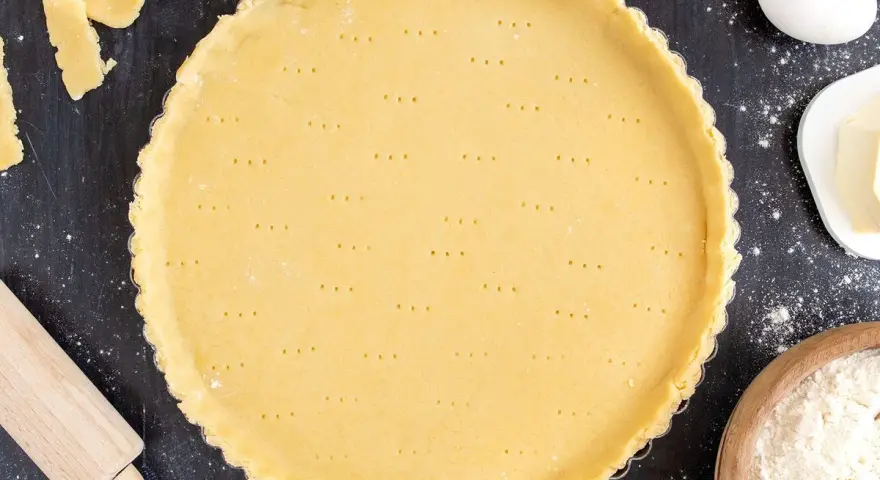 Ein vorbereiteter Mürbeteigboden in einer Tarteform, bereit für das 1 2 3 Teig Rezept. Zutaten wie Eier, Butter und Mehl sind daneben zu sehen.