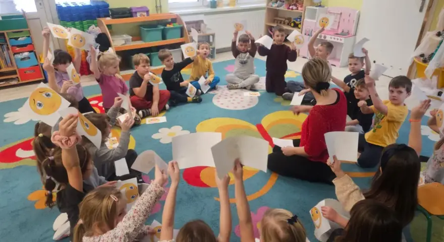 Wychowawca prowadzi zajęcia z dziećmi, które trzymają w rękach rysunki buziek wyrażających różne emocje.