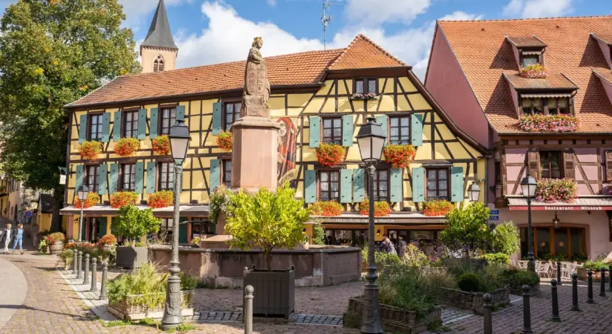 Ribeauvillé village médiéval : maisons à colombages colorées, statue, fontaine et fleurs.