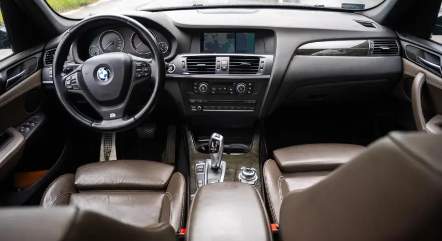 Wnętrze BMW X3 z brązową skórzaną tapicerką i drewnianymi wstawkami. Kierownica, deska rozdzielcza i nawigacja. Wiele osób szuka bmw x3 opinie, by poznać ten model.