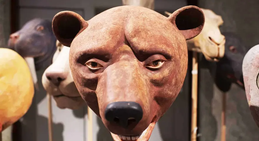 En brun björnmans ansikte, gjord av papier mache lim, med andra djurmasker i bakgrunden.