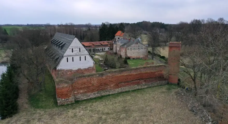Zamek Kiszewski, z ceglanymi murami i wieżą, otoczony zielenią i drzewami.