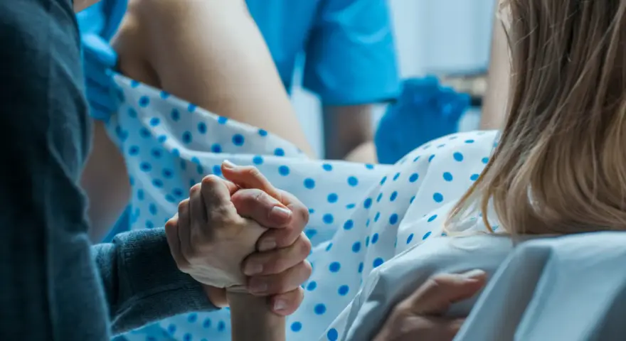 Wsparcie bliskiej osoby podczas porodu. Dłoń partnera trzyma dłoń rodzącej, która ma już widoczny czop śluzowy.