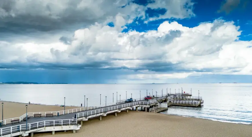 Długie molo w Brzeźnie wchodzi w spokojne morze. Na piaszczystej plaży widać kilka osób i samochody na molo.
