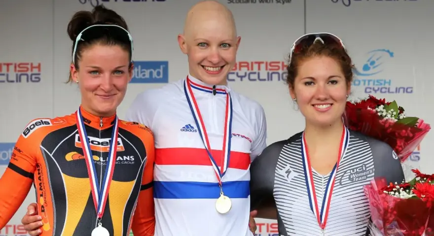 Joanna Rowsell, a bald cyclist in a white and red jersey, smiles on the podium with two other medalists.
