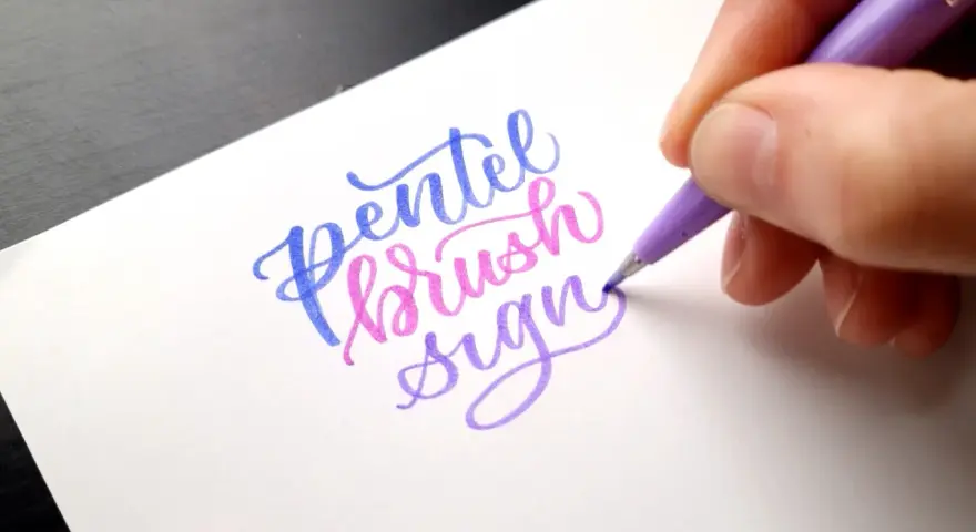 En hand skapar vacker brush lettering med en lila penna. Orden "Pentel brush sign" framträder i blått, rosa och lila.