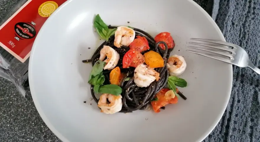 Czarny makaron z krewetkami, pomidorkami i bazylią na talerzu. Obok widoczna paczka spaghetti nero di seppia.