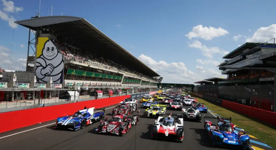 Una fila de coches de carreras listos para comenzar las 24 horas de Le Mans. El icónico muñeco de Michelin preside la tribuna.