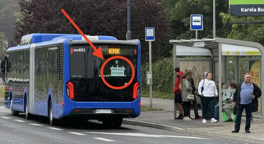 Niebieski autobus przegubowy z napisem "PRZEJAZD TESTOWY" na tyle, stoi przy przystanku autobusowym.