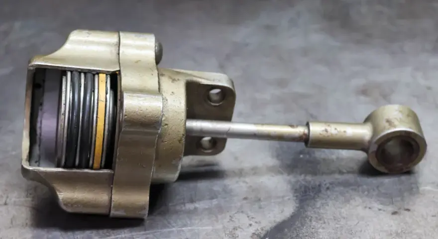 Metalowy element z tłoczyskiem i sprężynami, przypominający zawór wastegate. Objawy uszkodzenia mogą być widoczne na jego powierzchni.