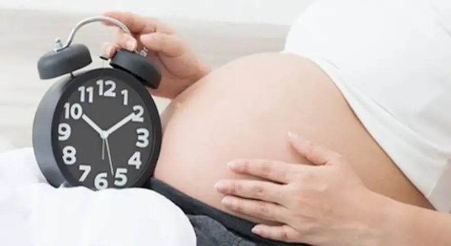 Zbliża się termin porodu. Kobieta trzyma dłoń na brzuchu obok budzika.