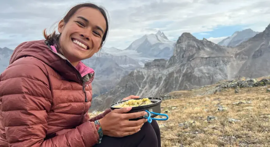 Sourire radieux d'une randonneuse savourant son repas en pleine montagne, avec des sommets enneigés en arrière-plan. Un moment de pur bonheur pour ce repas randonnée.