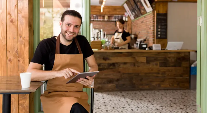 Hombre sonriente con delantal usa tablet en su cafetería, gestionando pedidos con un sistema erp restaurantes.