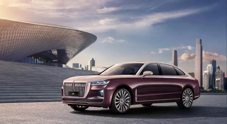Elegancki, bordowy Hongqi H9 z chromowanymi detalami stoi przed nowoczesnym budynkiem o falistej fasadzie, w tle miejski krajobraz.