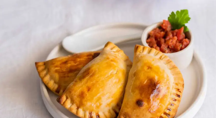 Tres empanadillas al horno doradas y crujientes, servidas en un plato blanco con un pequeño cuenco de salsa de tomate y una hoja de perejil.