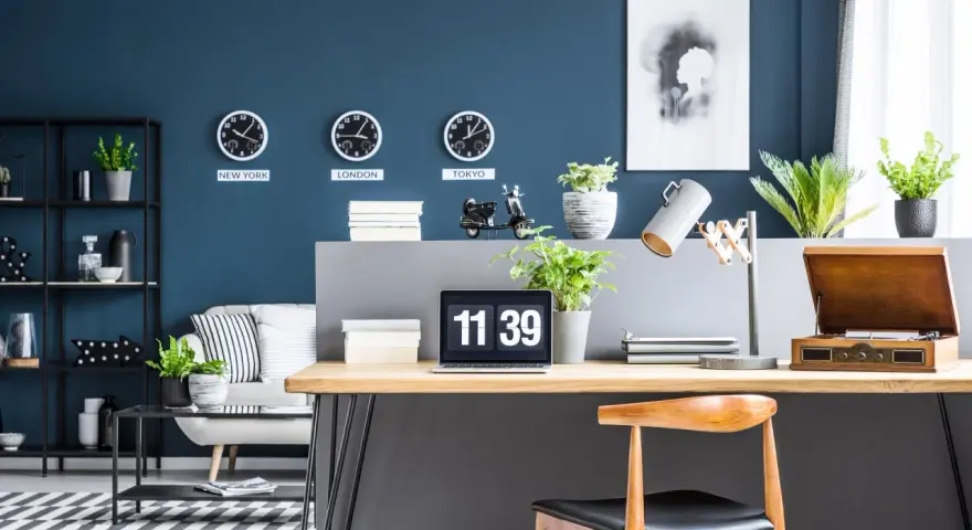 Biuro z zegarami pokazującymi czas w Nowym Jorku, Londynie i Tokio na ścianie w kolorze navy. Na biurku laptop, lampa i gramofon.