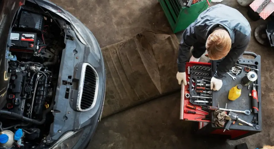Meccanico con guanti bianchi esamina un motore sbiellato, circondato da attrezzi e un'auto con il cofano aperto.