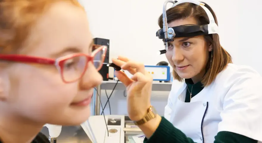 Lekarka bada ucho dziewczynki, która ma okulary. Badanie może pomóc zdiagnozować niedosłuch u dziecka.