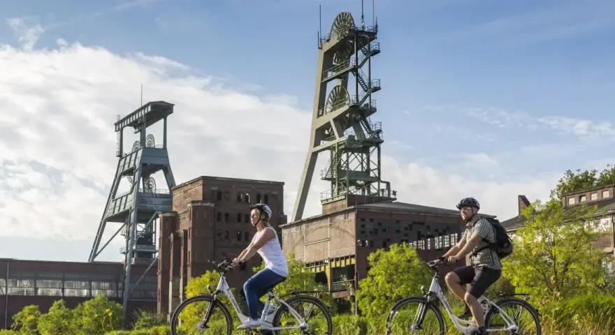 Zwei Radfahrer genießen die Landschaft am ehemaligen Bergwerk Ewald. Die imposanten Fördertürme stehen im Hintergrund.