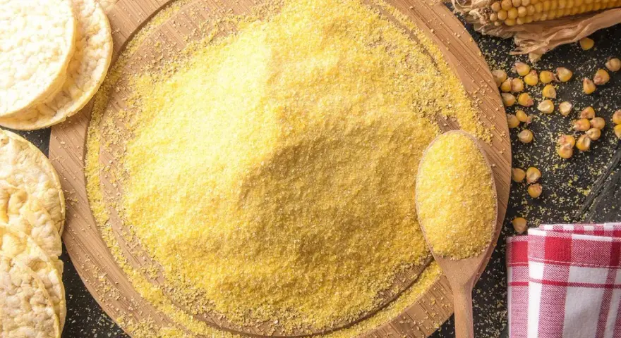 Stos mąki kukurydzianej na desce, obok łyżka z mąką, kukurydza i wafle.