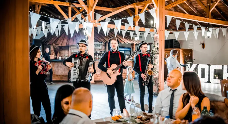 Zespół grający na weselu, z akordeonem, gitarą i saksofonem. W tle widać gości i dekoracje. Idealni do oprawy muzycznej imprez, znają teksty piosenek biesiadnych i weselnych.