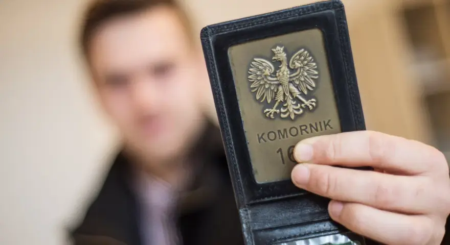 Komornik pokazuje legitymację z orłem. W tle niewyraźna postać mężczyzny, być może w związku z długiem osób prywatnych.