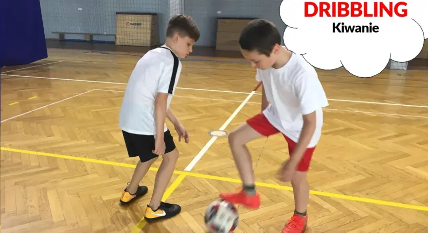 Dwóch chłopców ćwiczy drybling piłką nożną na sali gimnastycznej.