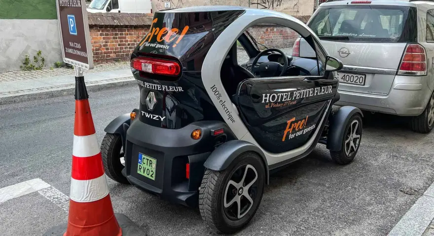 Czarny Renault Twizy z napisem "Free! for our guests" zaparkowany przed hotelem Petite Fleur. Obok znak parkingowy dla gości hotelowych.