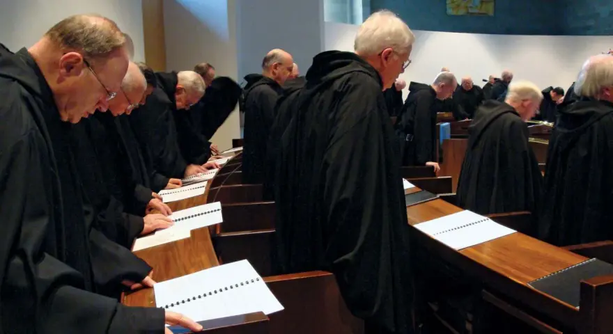 A group of Italian monks in black robes stand in a church, heads bowed over open prayer books.