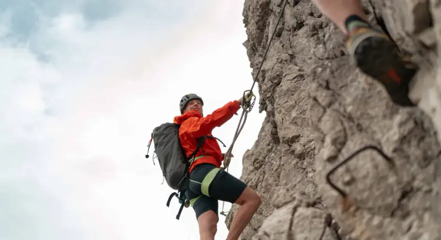 Kletterin im orangefarbenen Anzug sichert sich am Drahtseil des Alpspitz Klettersteigs. Ein spannendes Abenteuer in den Bergen.