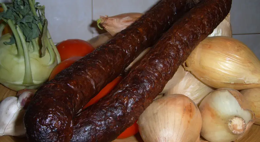Wędzona kiełbasa z dzika na talerzu z warzywami: cebulą, pomidorami, kalarepą i czosnkiem.