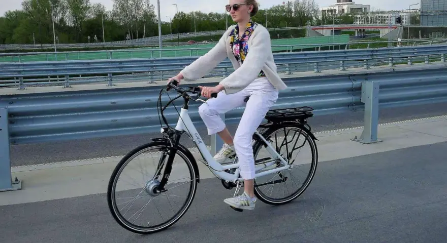 Kobieta w białych spodniach i sweterku jedzie na rowerze elektrycznym. W tle widać most i budynki.