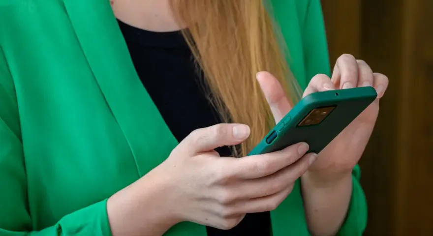 Kobieta w zielonej marynarce trzyma telefon. Szuka informacji, gdzie jest schowek w telefonie, aby ukryć ważne dane.