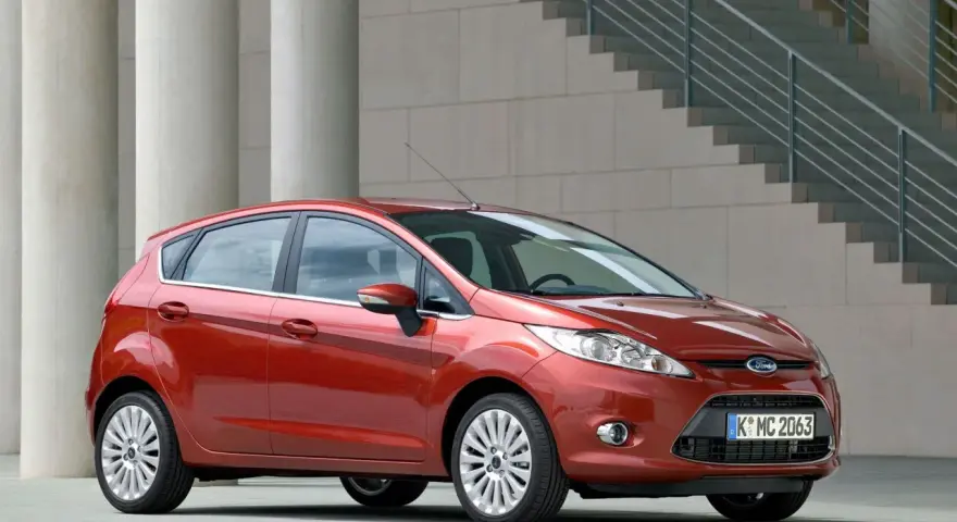 Czerwony Ford Fiesta MK6 zaparkowany przed nowoczesnym budynkiem z betonowymi kolumnami i schodami.