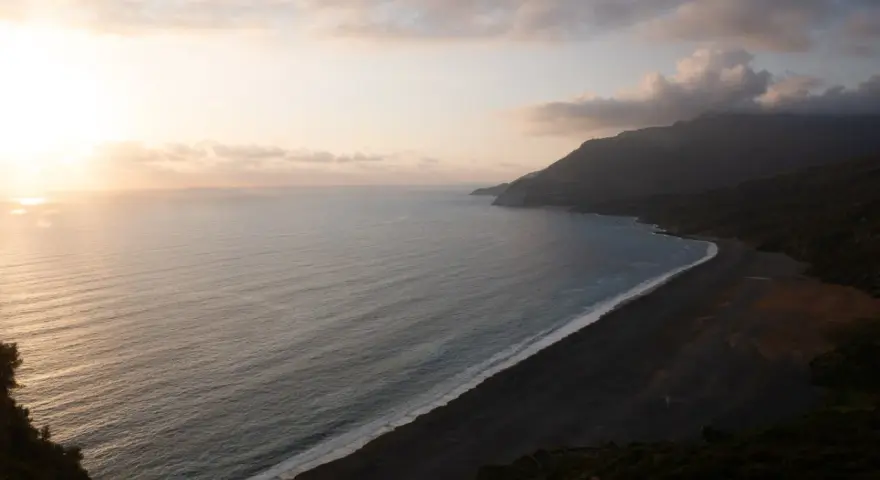 Vue aérienne d'une plage de sable noir bordée par une mer calme et des montagnes embrumées. Ces photos de plage capturent la sérénité d'un paysage côtier au coucher du soleil.