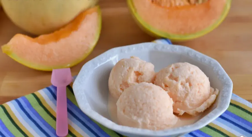 Tre palline di cremoso gelato al melone in una ciotola bianca, con fette di melone fresco sullo sfondo.