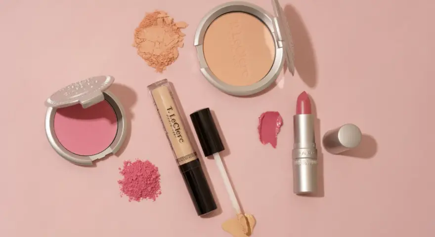 Un assortiment de maquillage rose et doré : blush, poudre, fond de teint, rouge à lèvres et fard à paupières sur fond rose.