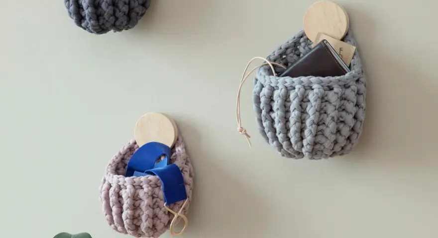 Three crocheted wall pockets in grey and pink, one holding a wallet, another blue ribbon, on a light wall.