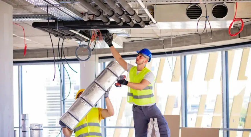 Dwóch pracowników montuje systemy HVAC, instalując izolację na rurach. To kluczowy element zapewniający komfort cieplny.